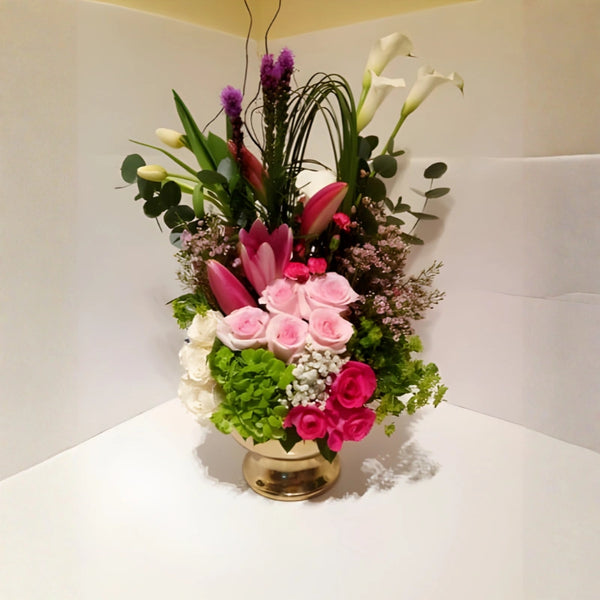Floral arrangement with pink roses, green leaves, and other flowers in a gold vase on a white surface.